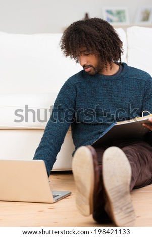 Young African American man studying at home sitting with his back to the sofa on the floor in the living room looking up information on his laptop computer