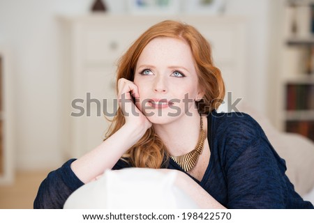 Beautiful young woman sitting thinking leaning over the back of a comfortable sofa staring up into the air with a contemplative expression