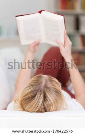 View from the top of the head of a blond woman lying on her back on a sofa at home reading a book