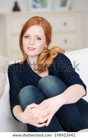 Beautiful pensive woman relaxing at home sitting on the couch with her knees up and hands clasped around them looking thoughtfully at the camera