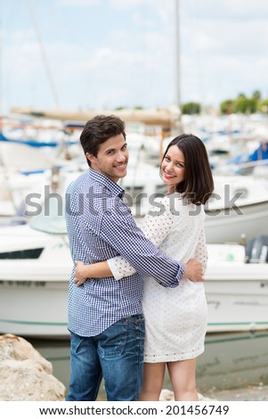 Attractive young couple standing arm in arm overlooking a marine small boat harbour turning back to smile at the camera