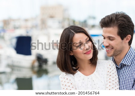 Attractive happy young couple standing close together with the woman turning to look at her husband as he laughs in amusement, with copyspace on a blurred urban scene