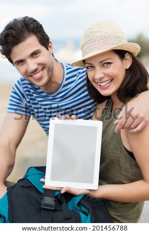 Stylish affectionate young couple sitting at the beach displaying a blank tablet computer above a backpack and smiling at the camera with beaming happy smiles