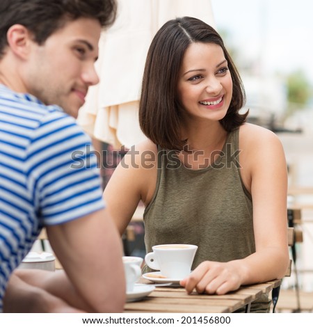 Young couple having coffee outdoors sitting at an open-air cafeteria turning to watch something on the right, focus to the smiling beautiful woman