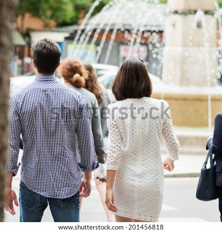 Young couple walking with their backs to the camera in an urban street with a crowd of people as they approach a fountain with cascading jets of water