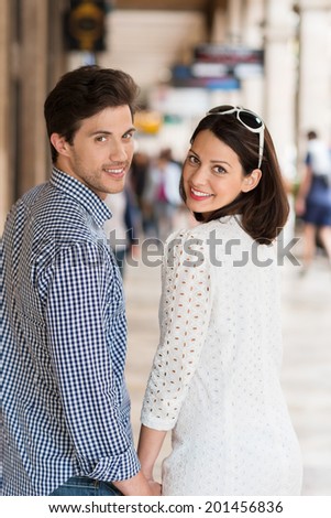 Stylish young couple turning to look back at the camera as they walk hand in hand along an urban street