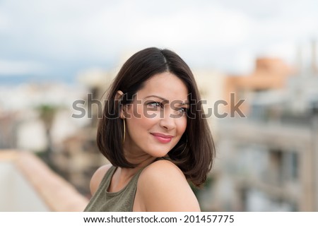 Smiling beautiful woman looking back over her shoulder at the view as she leans against a wall on a balcony overlooking a town