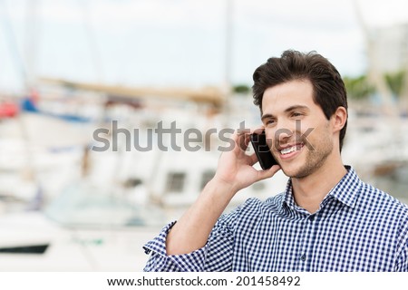 Handsome unshaven young man chatting on a mobile phone smiling at the conversation against a small boat harbour background