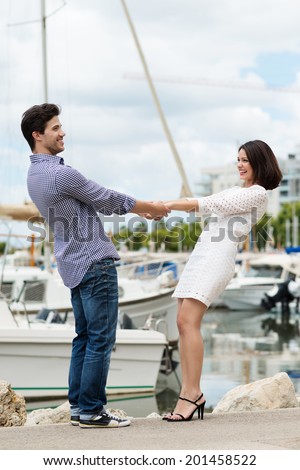 Energetic young couple on a quay overlooking boats in a harbour laughing and holding hands and leaning back pulling against each other