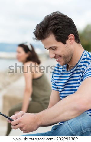 Young man reading an sms on his mobile phone and smiling at the content as he sits on a wall overlooking the ocean