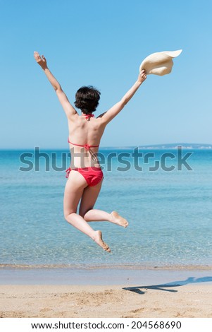 Young woman in a red bikini leaping for joy on a tropical beach raising her arms in the air as she holds onto her her straw sunhat, view from behind overlooking a calm blue ocean