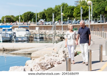 Happy middle-aged couple walking hand in hand past a small boat marina at the coast as they enjoy an active day in the summer sun
