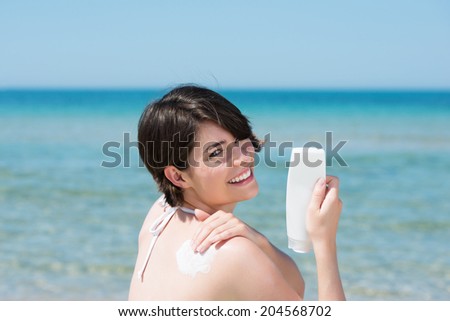 Beautiful young woman on the beach applying suntan lotion or cream to her back from a plastic bottle turning to smile happily at the camera against a calm blue ocean backdrop