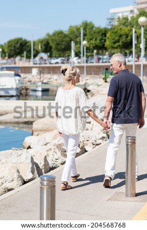 Middle aged couple strolling hand in hand past a small boat harbour on a hot summer day with their backs to the camera