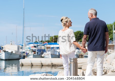 Mature couple enjoying a day at the coast walking away from the camera hand in hand past a small boat harbour