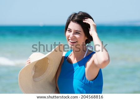 Beautiful laughing woman at the seaside standing in front of the ocean holding a wide brimmed straw sunhat with her hand to her short brunette hair