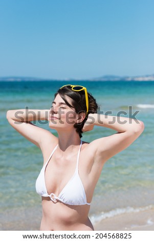 Beautiful young woman in a bikini standing on a tropical beach with her hands clasped behind her neck and sunglasses perched on her head celebrating the sunshine with a smile of bliss