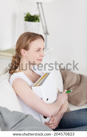 Attractive young woman relaxing on a sofa at home clutching a large binder to her chest as she stares thoughtfully into the distance