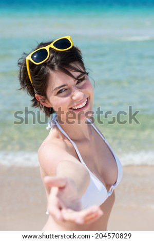 Beautiful woman in a bikini holding out an inviting hand towards the camera with a charming friendly smile as she urges you to join her on the sunny tropical beach