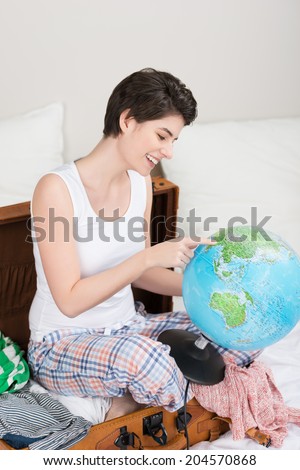 Smiling beautiful woman sitting on a half packed suitcase pointing to a globe selecting a destination fro her summer vacation