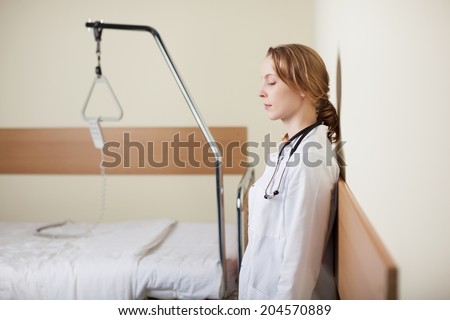Young female doctor standing with closed eyes leaning against the wall in a ward alongside an empty hospital bed as she takes a moment to gather herself together