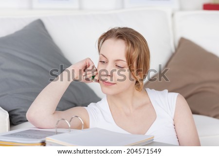 Beautiful young woman studying at home sitting leaning back against the sofa reading her notes in a large binder