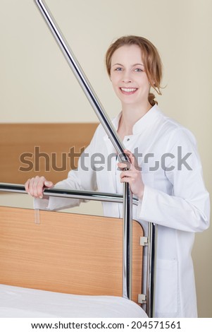 Beautiful happy young female doctor or nurse in a white lab coat standing holding the rail on a hospital bed smiling at the camera