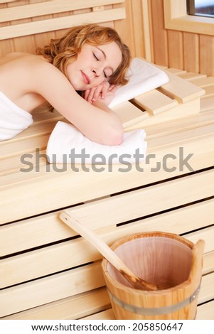 Woman sleeping in a sauna bath lying on her stomach on a wooden bench with her eyes closed with the water bucket and ladle in the foreground