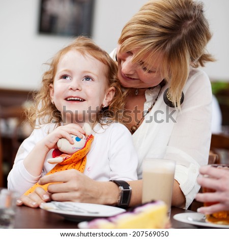 Cute little girl holding toy with mother sitting at table in restaurant