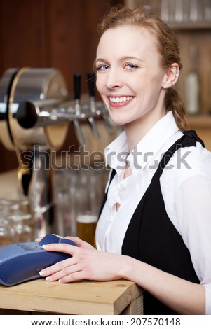 Smiling waitress in a bar standing behind the counter with her hands resting on a portable credit card machine for payment for services