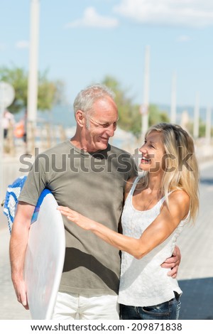 Happy fit tanned middle-aged couple going surfing walking along a sunny street arm in arm carrying a surfboard