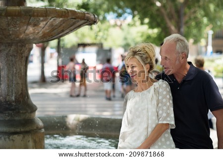 Laughing mature standing couple alongside a fountain in a shady urban square watching the water cascading down
