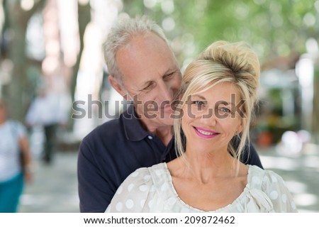 Loving mature couple in an intimate embrace with the husbands nuzzling his wife from behind as she looks at the camera with a beautiful smile, outdoors urban background