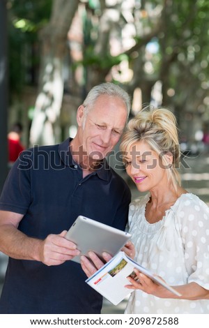 Couple checking information on a tablet computer as they reference a travel brochure while on summer vacation in a tree lined urban street