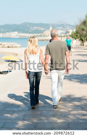 Middle-aged couple walking on a seafront promenade walking away from the camera hand in hand in the hot summer sunshine