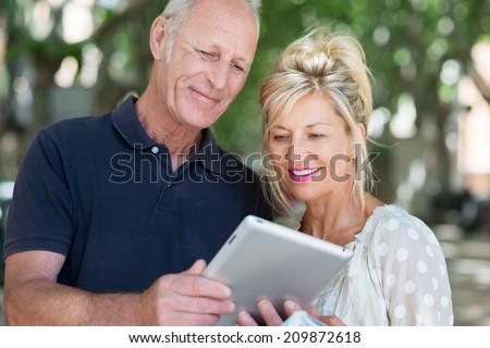 Attractive mature couple standing in a tree lined urban street reading information on a tablet computer with a smile