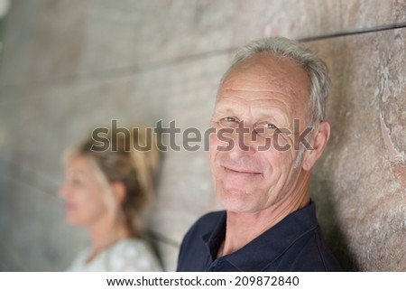 Smiling confident grey-haired senior man leaning against a receding wall turning his head to look at the camera with a charming smile