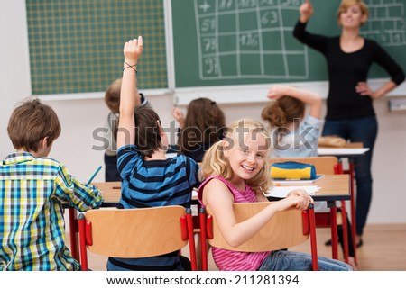 Beautiful little blond girl sitting in class turning to smile at the camera as the teacher takes questions from the rest of her classmates