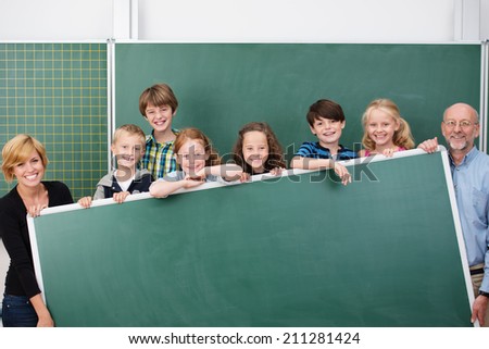Happy school team of young students and teachers standing behind a large black chalkboard with copyspace smiling happily at the camera