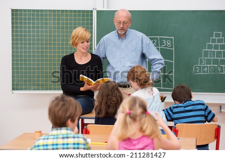 Teacher with his young attractive female teaching assistant standing in front of a class of small girls and boys as they discuss something from a textbook