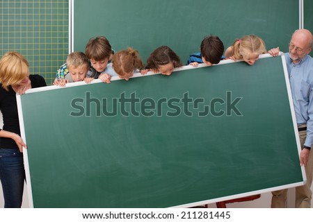 Young students and their two teachers standing holding a blank blackboard all peering over the top to see what will be written there
