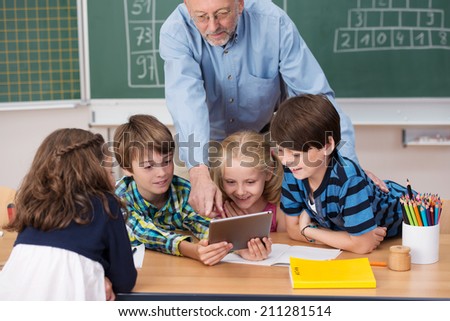 Smiling young children using a tablet in class as they cluster around a young girl sitting at a desk with a male teacher behind pointing out something on the screen