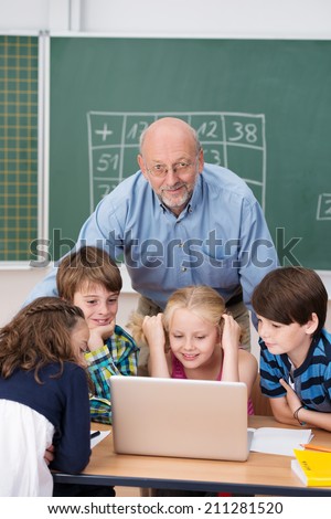 Smiling elderly male teacher leaning over his young students clustered around a laptop computer on the desk