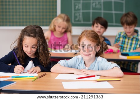Smiling pretty young redhead girl in class sharing a desk with a young classmate looking up from her notes to give the camera a beaming smile