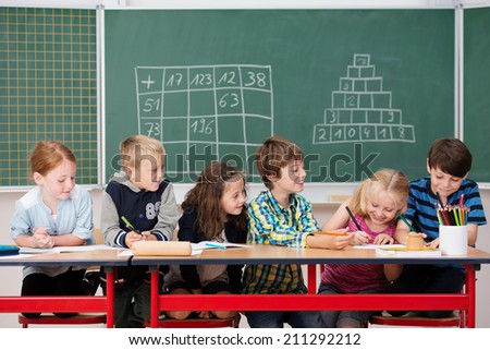 Group of happy children at work in the classroom sitting working on their projects at a long table in front of a blackboard