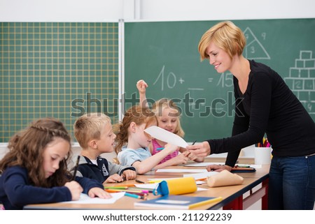 Attractive young female teacher checking a young boys work as he sits working on a project with other students at a long table
