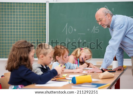 Elderly male teacher in primary school leaning over a long desk chatting to a group of young boys and girls with the blackboard in the background
