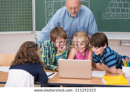 Young school kids in class grouped around a laptop computer on a desk with a male teacher leaning over watching them work
