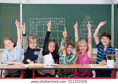 Enthusiastic group of young kids in class sitting in a row at their desk raising their hands in the air to show the know the answer to a question