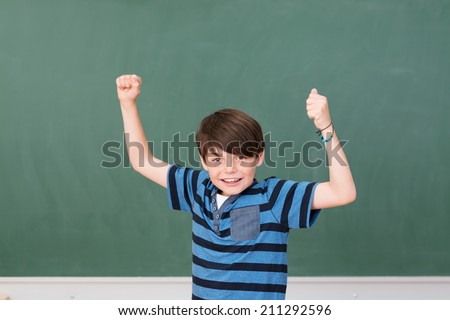 Handsome young schoolboy celebrating in the classroom cheering and punching his fists in the air